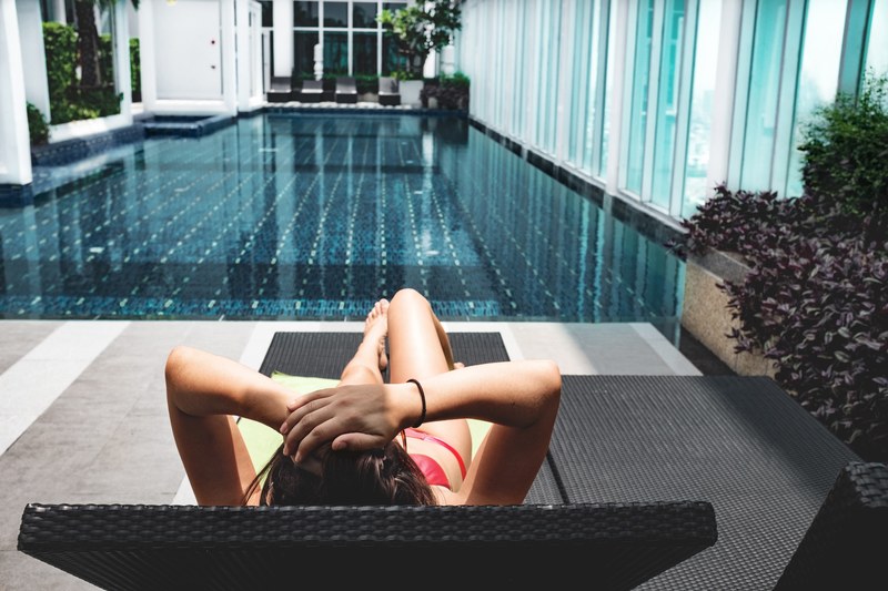 a woman in a bikini lays on a lounge chair facing a pool, summer resplendence 4k