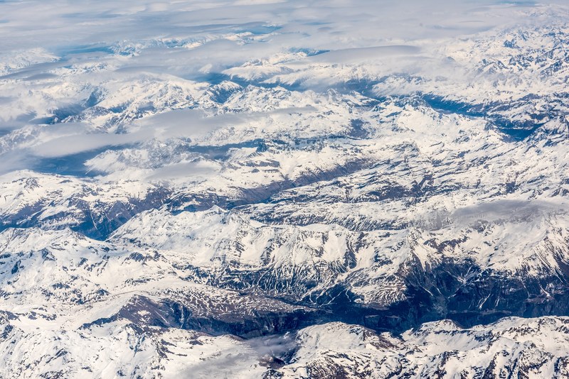 an aerial shot of a snowy mountain range under patchy clouds, birds eye view of snowy mountains 4k