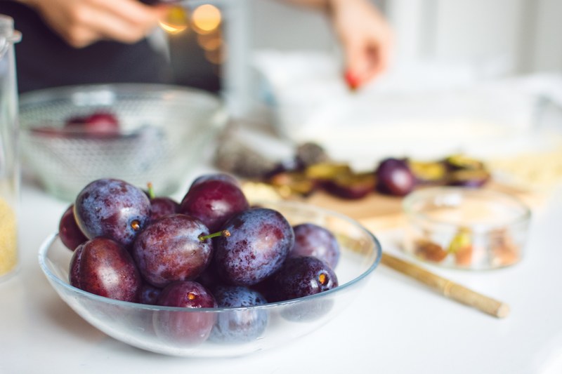 glass bowl of purple plums on counter in front of other fruits and a persons hands, baking plum cake 4k