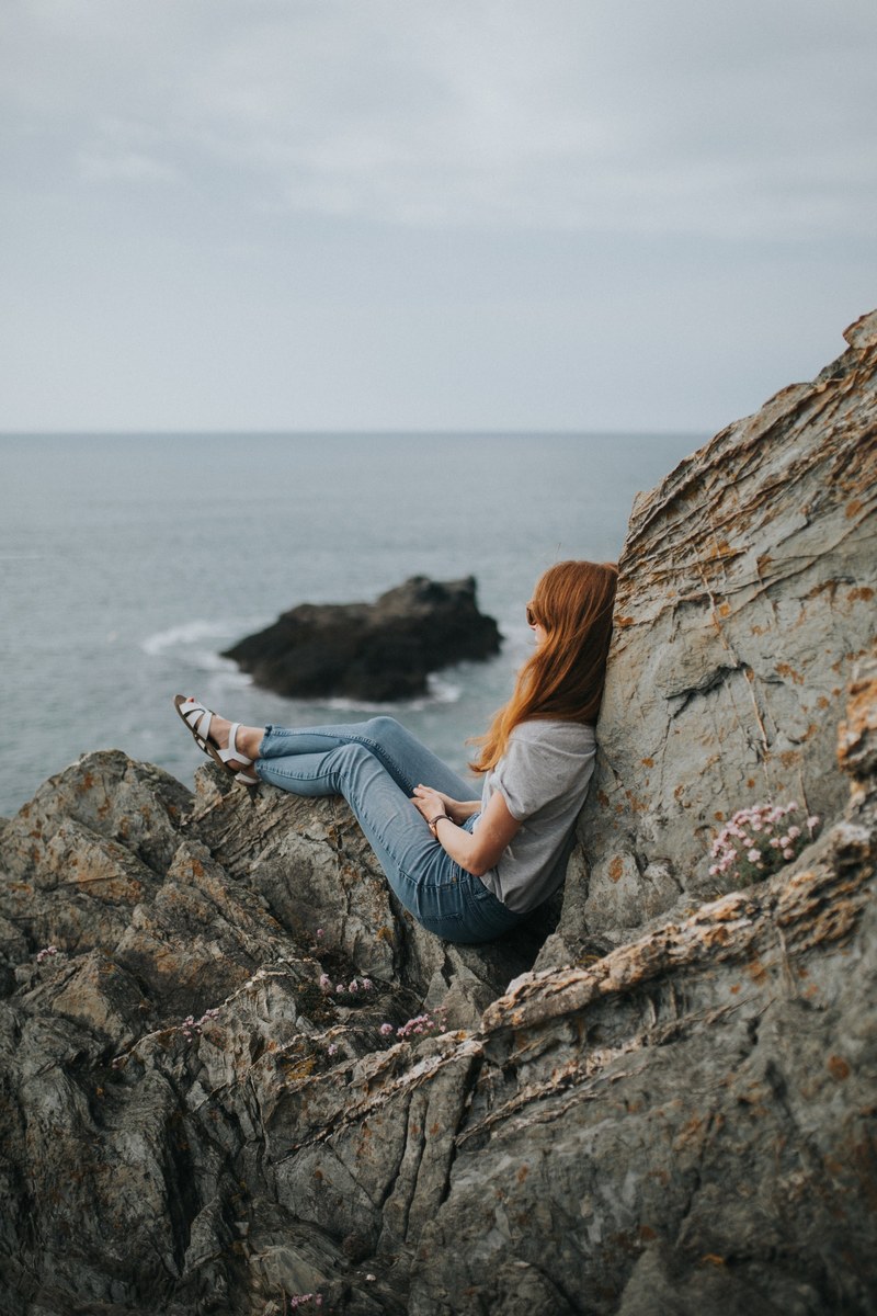 woman in jeans and shirt sitting on coastline rocks at fistral beach, woman sitting on the rocks 4k