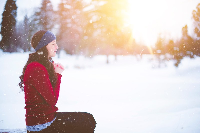 a woman wearing a blue headband and red sweatshirt holding her hands together in prayer while sitting outside on a snowy day, woman praying during winter 4k
