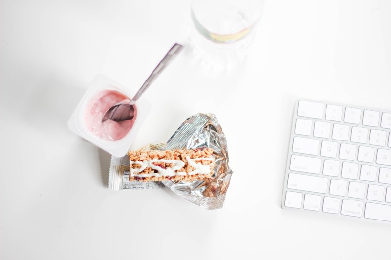 a spoon inside a small yogurt cup next to a granola bar and a mac keyboard, _snacking 4k
