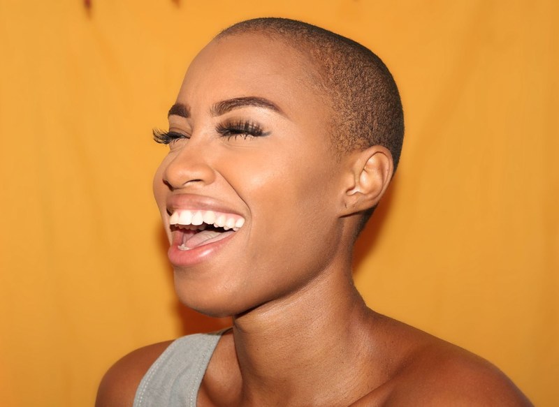 a woman with a shaved head smiles broadly against a yellow background, all smiles 4k