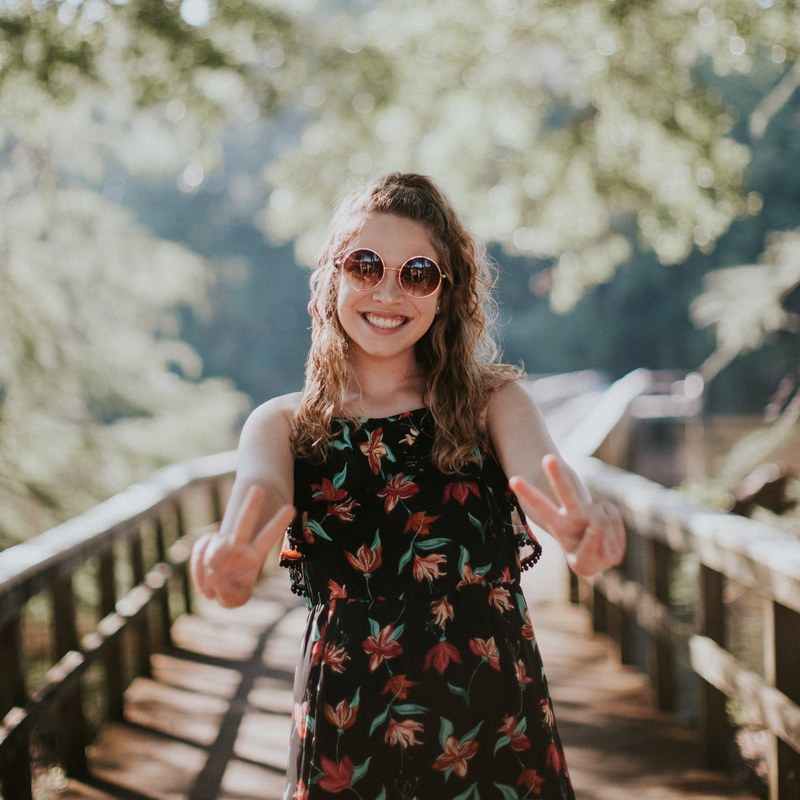 a smiling woman in sunglasses and a dress stands on a pier holding up peace signs, girl with sunglasses on a pier 4k