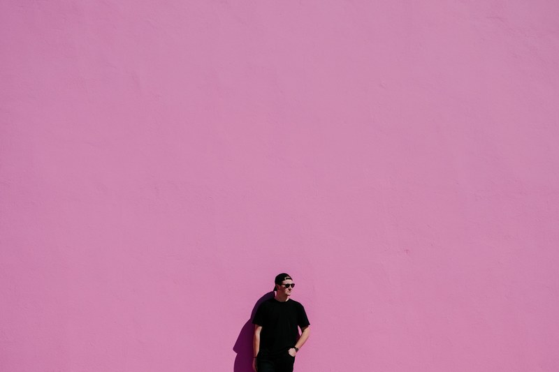 a man in a backwards baseball cap black t shirt and watch stands against a pink wall, man against pink wall 4k