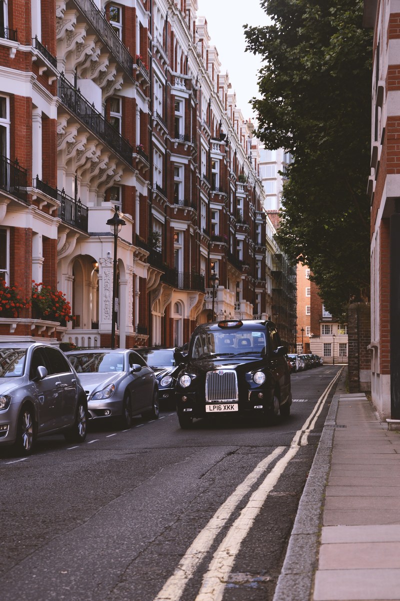 vintage black car driving down a street with brick apartment complexes and other cars along either side, cab in london 4k