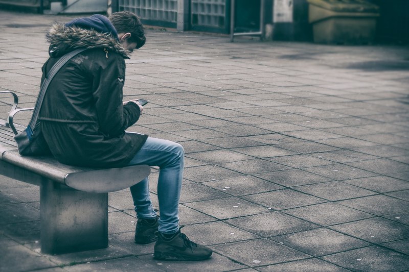 a person in a coat sits on a city bench looking down at their phone, person looks down at phone 4k