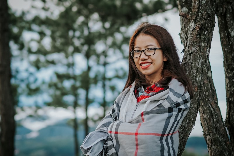 a smiling woman in glasses and a plaid cape leans against a tree in dalat, when in dalat 4k