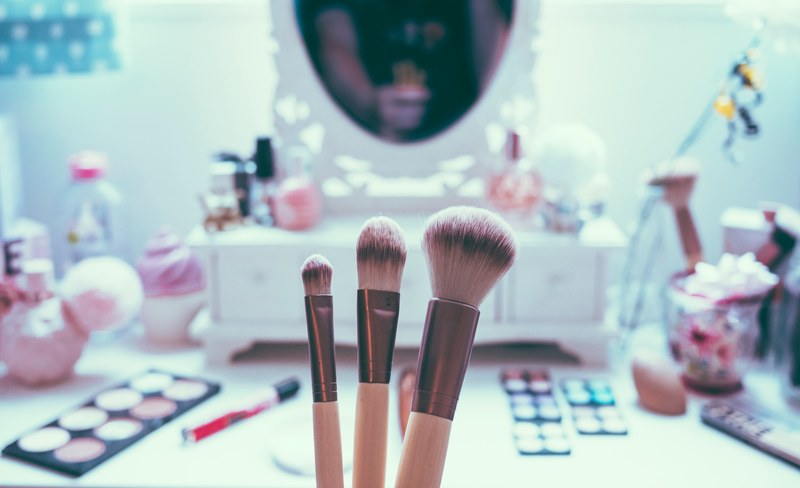 woman holding up three makeup brushes in front of a make up mirror with blush lipstick brushes and lights surrounding it, makeup brushes 4k