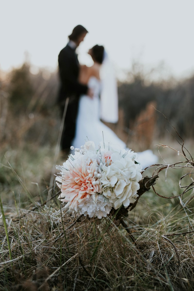 a bouquet sits in tall grass while a married couple embraces in the fuzzy background, wedding evening 4k