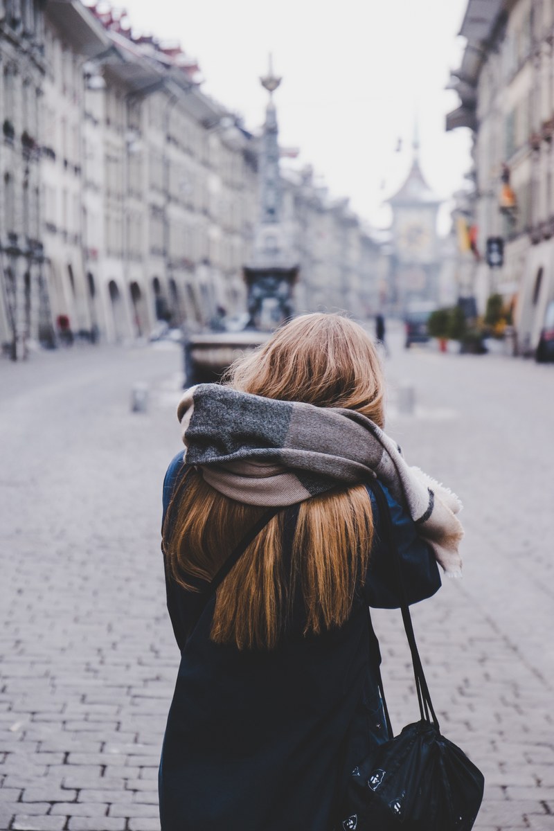 a woman in a scarf faces away down a gray brick street toward a city, the unknown girl 4k