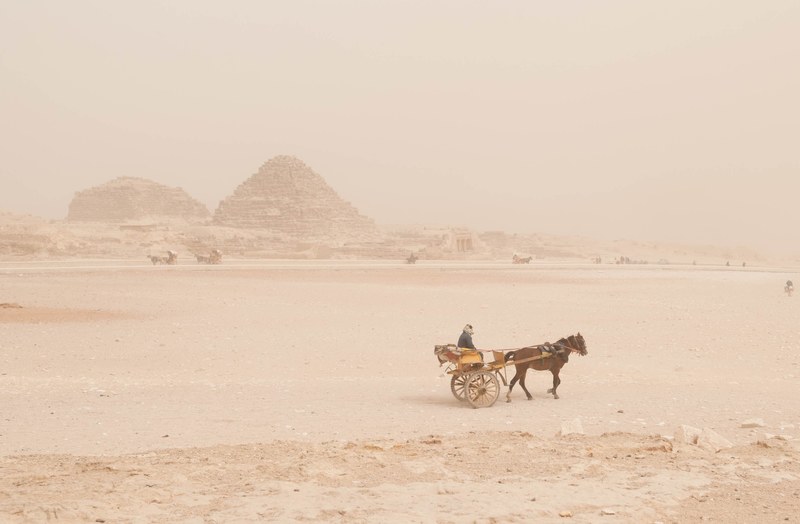 a woman sits on a small cart as a brown horse pulls it through the rocky desert, sandstorm is coming 4k