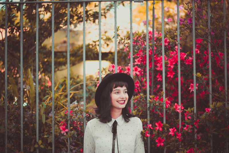 a young woman in a hat smiling while standing next to flower bushes, woman in a hat by red flowers 4k