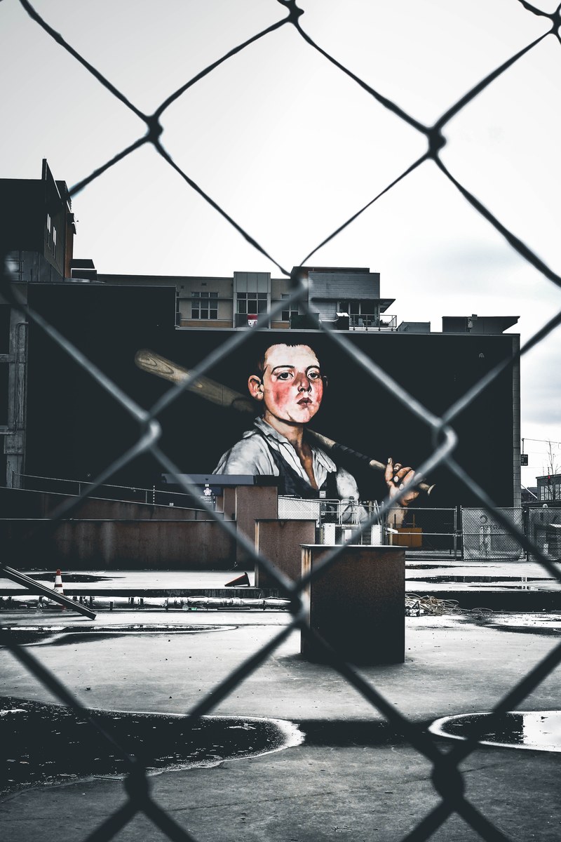 the view of art of man with baseball bat from a chain fence of an urban building, puddles 4k