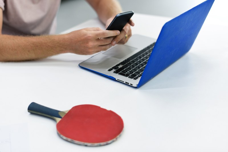 a person using their smartphone with their hands hovering over their blue laptop and a red table tennis paddle nearby, _blue laptop red tennis paddle 4k