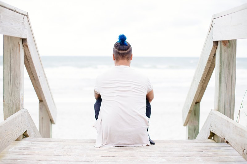 a man with a blue dyed bun sitting on wooden stairs near the sea, lonely man near the sea 4k