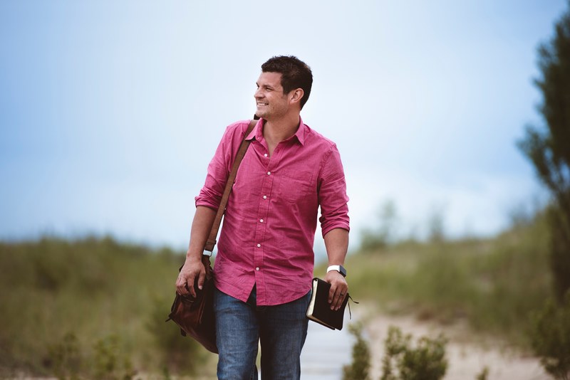 a man in a pink shirt with a leather bag and a book walking on a dirt path, man holding leather bound book 4k