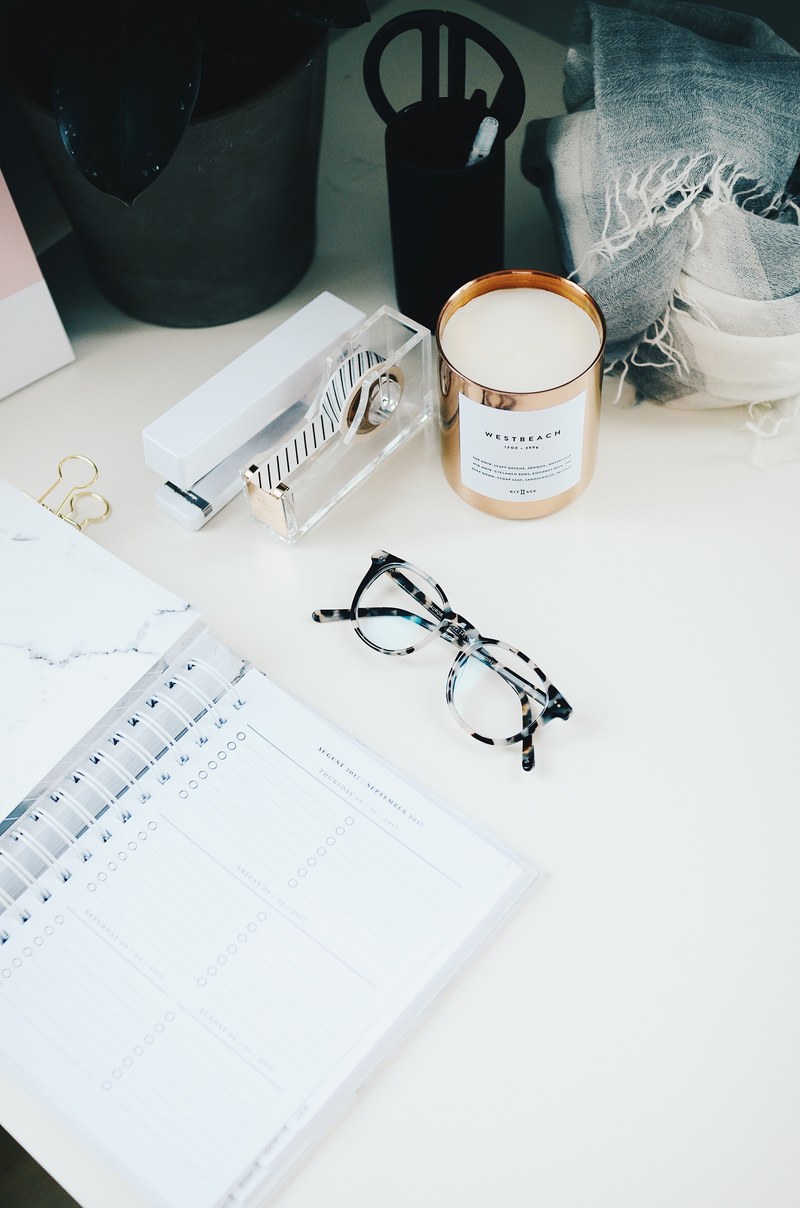 a pair of glasses next to an open planner and a scented candle on a white surface, saturdays desk 4k