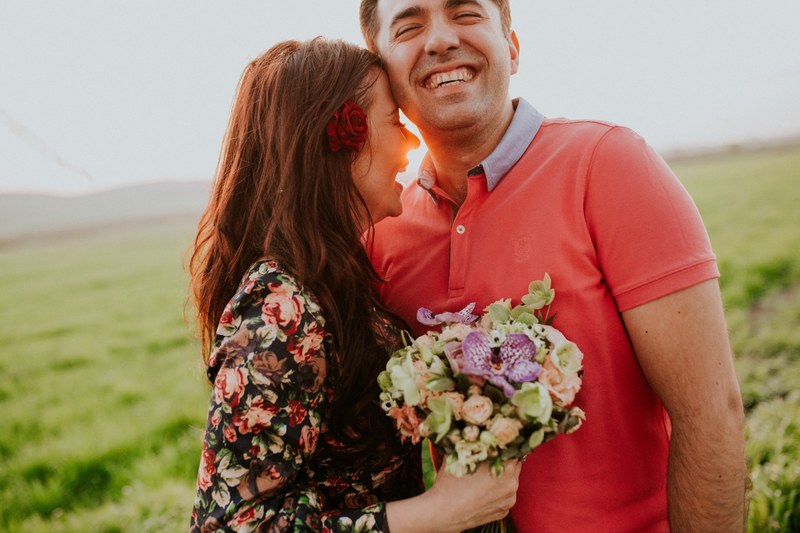 a man and a woman laughing in a sunny field she holds a bouquet of flowers, _36 4k