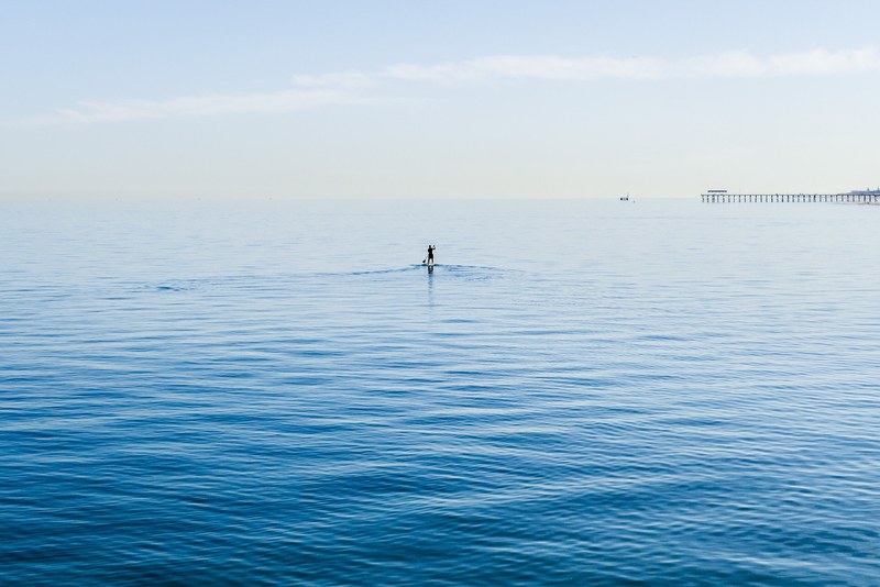 person on a paddle board in the ocean with the pier in the distance at henley beach, stand up reach out 4k