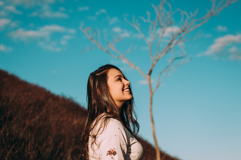 a woman smiles into the distance on a grassy hillside before a tree and blue sky, im a dreamer_ 4k