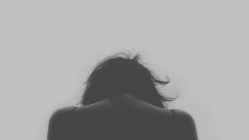 a black and white rear shot of the back of a womans head and back with her head bowed down in sadness, female head from behind 4k