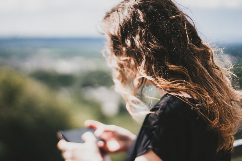 a woman with brown curly hair looks down to use her cell phone outdoors, woman looking at cell phone 4k
