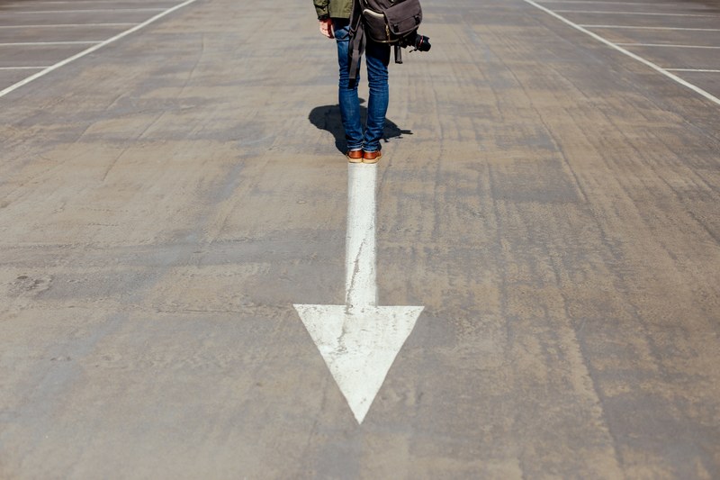 a low shot of a person with a backpack and a camera standing on a white arrow on asphalt, man on arrow 4k