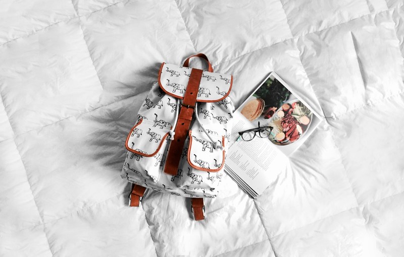 a white backpack on a bed mattress next to a cookbook and a pair of glasses, backpack and cookbook on a bed 4k