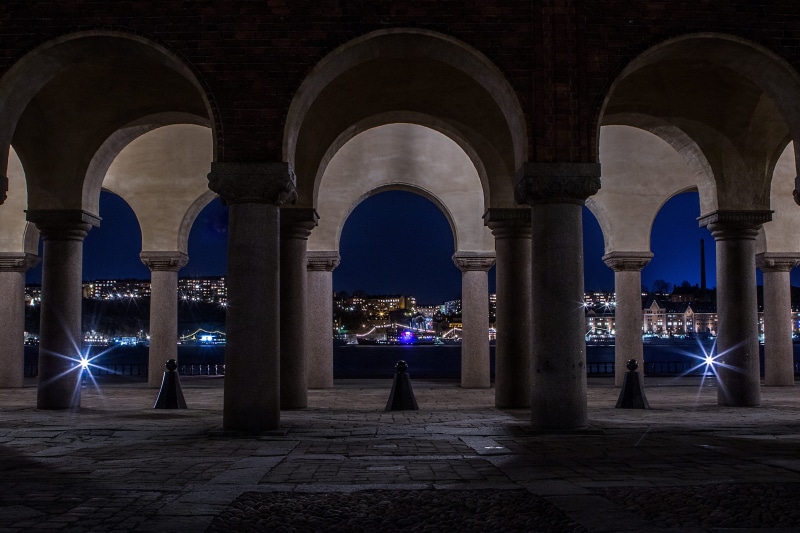 building exterior, illuminated, walkway, column, town, pillar, long expo, outdoors, corridor, light, men, architecture, floor, real people, crypt, architectural column