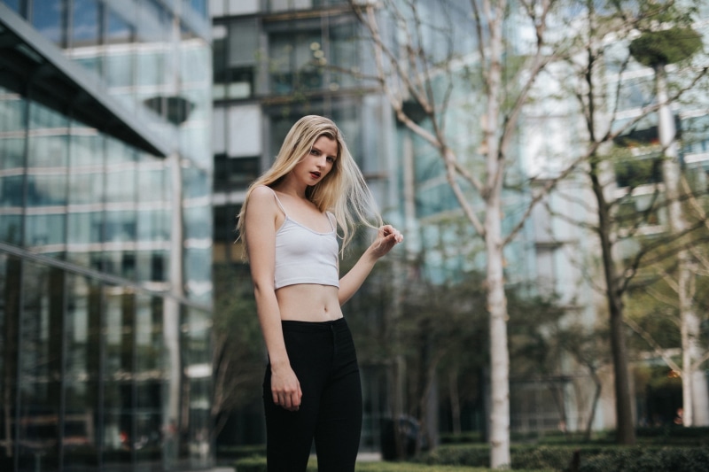 standing, built structure, day, long hair, red lipstick, hairstyle, one person, women outdoors, blond hair, touching hair, building, portrait, street, young adult