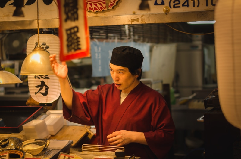 man, lamp, monk, apparel, building, indoors, town, occupation, arcade game machine, retail, working, kyoto prefecture, small business, shop, hat, clothing