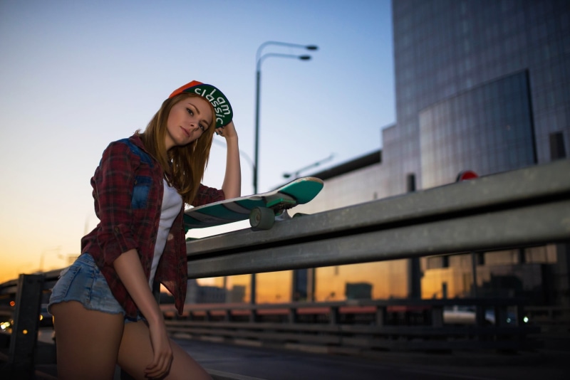 jean shorts, leaning, city, rails, fashion, sunset, real people, 2K, woman, architecture, casual clothing, red, Public, sport, skateboard, hairstyle, built structure