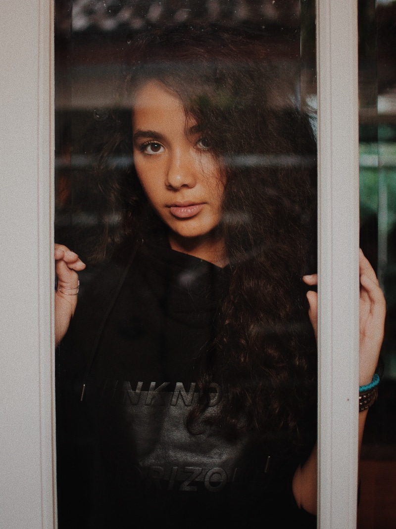 glass - material, hair, leaning, holding, black, looking, hoodie, real people, contemplation, pullover, front view, reflection, indoors, glass, door, waiting