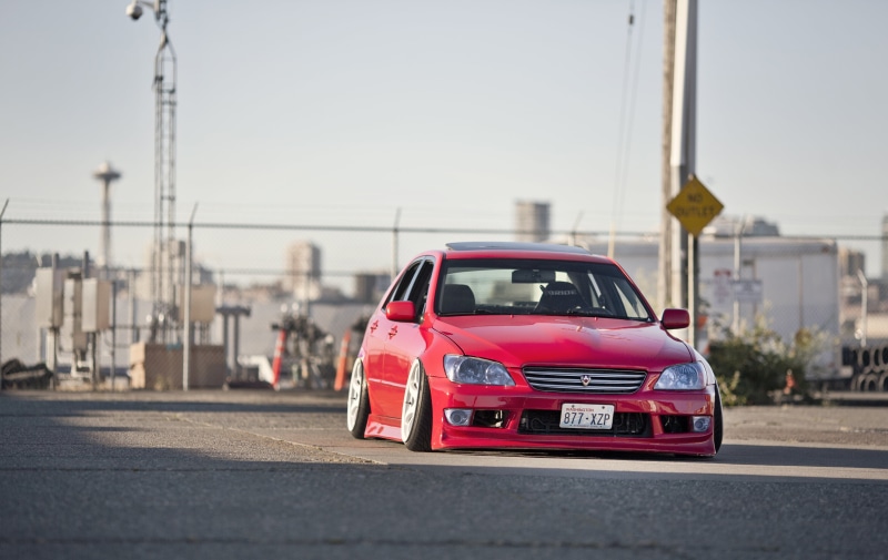 building exterior, red, clear sky, no people, sunlight, motion, Lexus, nature, motor vehicle, focus on foreground, mode of transportation, 4K, built structure