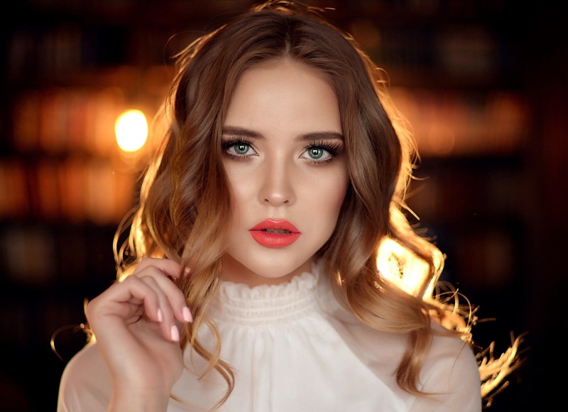 looking at viewer, bokeh, glamour, women, green eyes, one person, curly hair, touching hair, red lipstick, hairstyle, white clothing, human body part, brunette