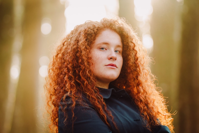 black, curly hair, frizzy, focus on foreground, style, outdoors, pose, beautiful woman, facial expression, lens flare, woman, redhead, one person, contemplation