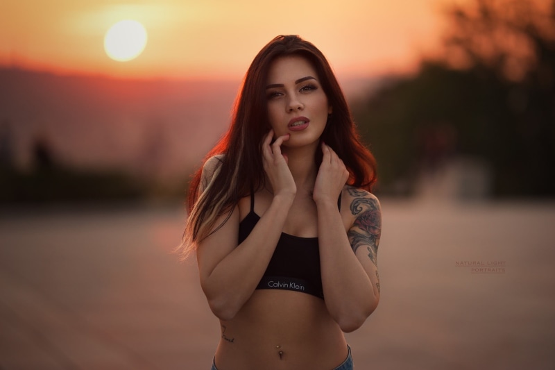 lips, sun, open mouth, 1080P, hips, pierced navel, black top, strap, bokeh, top, touching face, looking at camera, face, portrait, brown eyes, depth of field