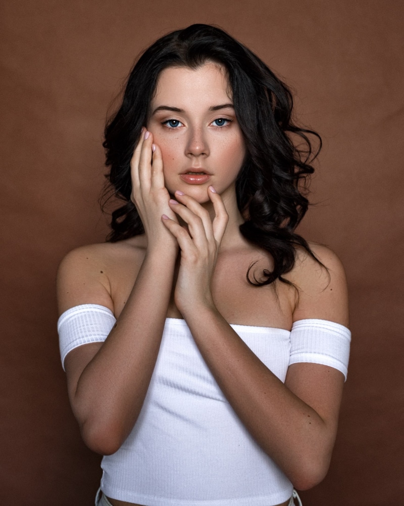 white tops, looking at viewer, model, women, Max Pyzhik, indoors, Daria Delishes, brunette, typographic portraits, touching face, bare shoulders, brown background