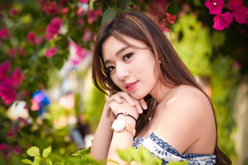 brunette, face, bracelets, outdoors, bokeh, brown eyes, looking at viewer, Kiki Hsieh, pink lipstick, 2K, depth of field, flowers, plants, long hair, pearl earrings