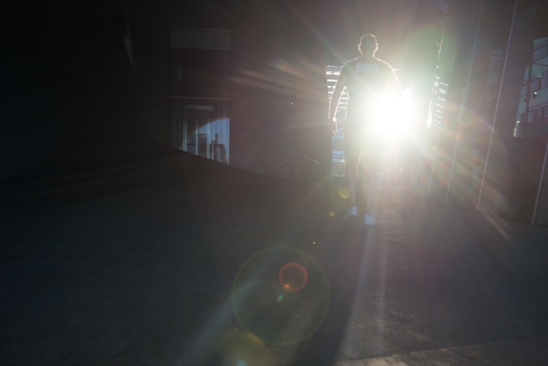lens flare, walking, real people, building exterior, human, one person, standing, 2K, person, full length, shadow, transportation, man, men, street, illuminated