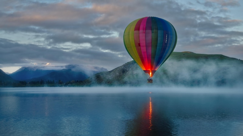 tranquil scene, mid-air, mountain, reflection, hot air balloon, multi colored, nature, lake, tranquility, flying, no people, morning, beauty in nature