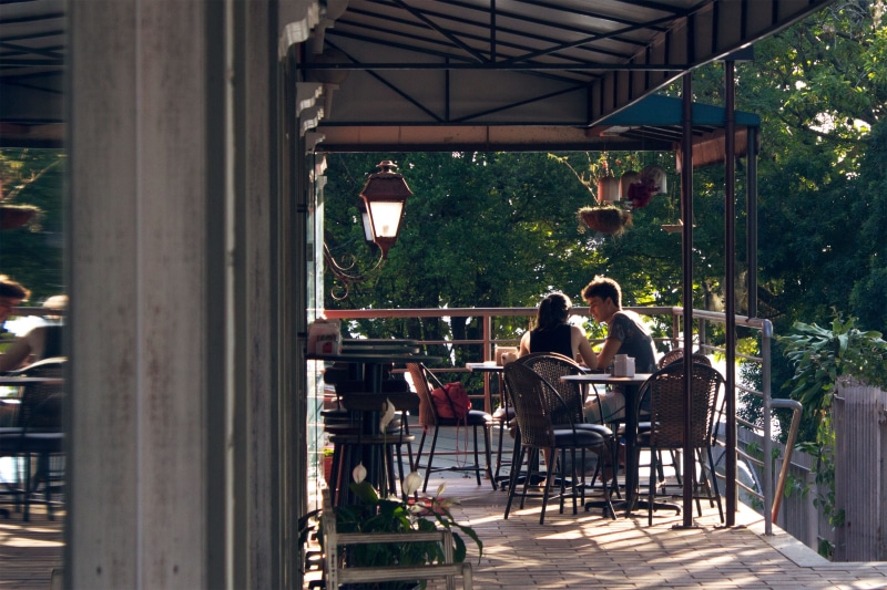 near, chairs, 2K, building, woman, patio, architecture, table, outdoors, people, women, business, plant, coffee shop, day, cafe, sitting, daylight, lamppost