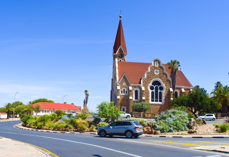 sky, mode of transportation, historically, religion, tree, road, landmark, german, belief, motor vehicle, old, blue, clock, windhoek, church, transportation