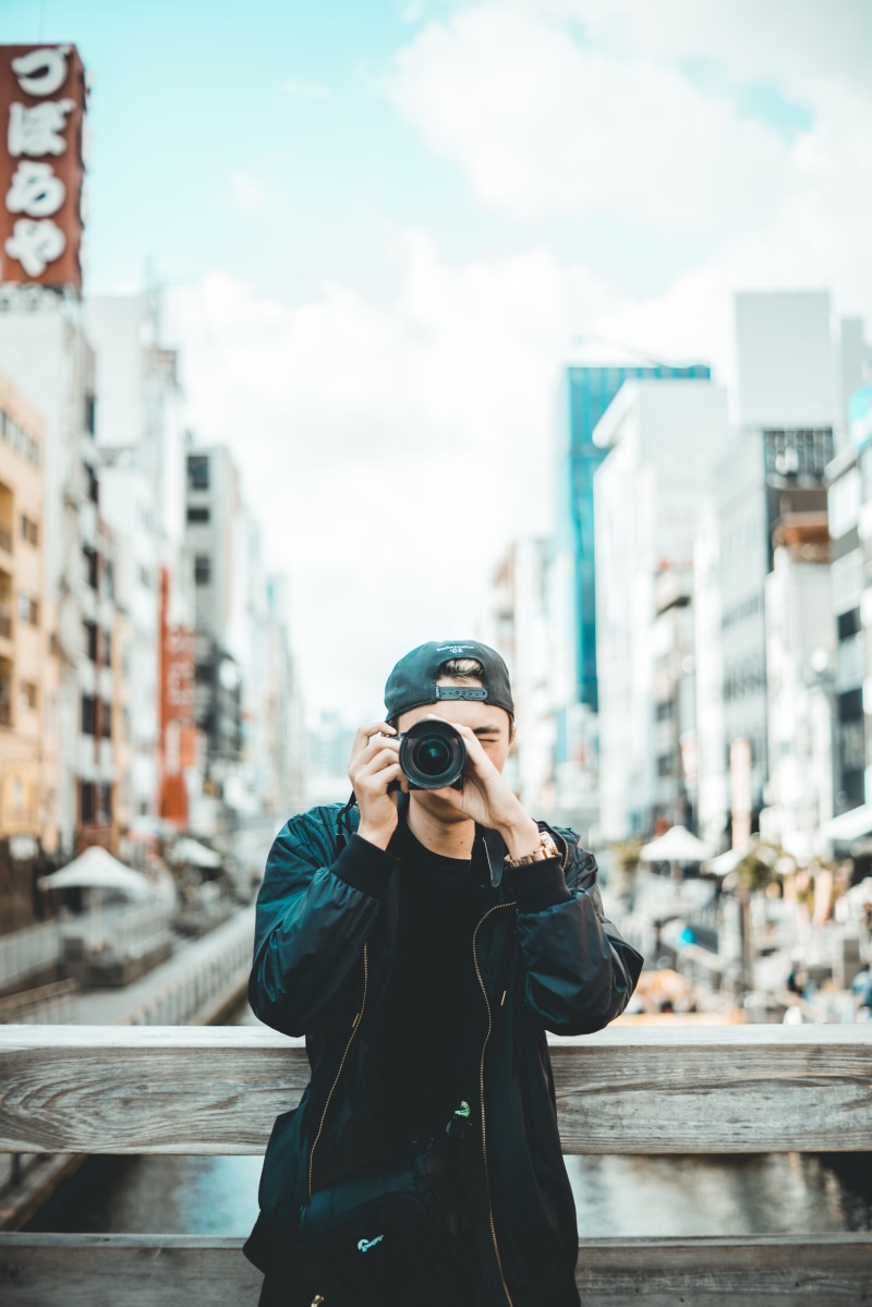 technology, camera - photographic equipment, male, near, building exterior, taking photo, streetscape, outdoors, daytime, focus on foreground, cap, standing