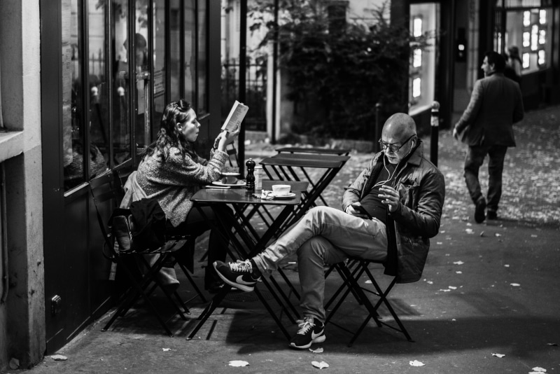 street, casual clothing, grayscale, 4K, sidewalk, holding, women, seat, focus on foreground, store, people, social issues, chairs, city, pavement, reading
