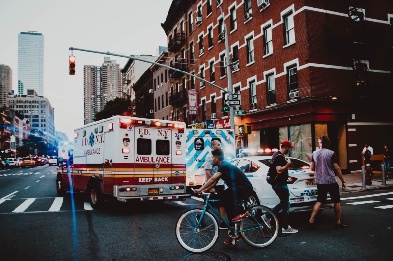 mode of transportation, street, sign, riding, life, urban, ambulance, incidental people, women, sky, bicycle, building exterior, man, near, motor vehicle