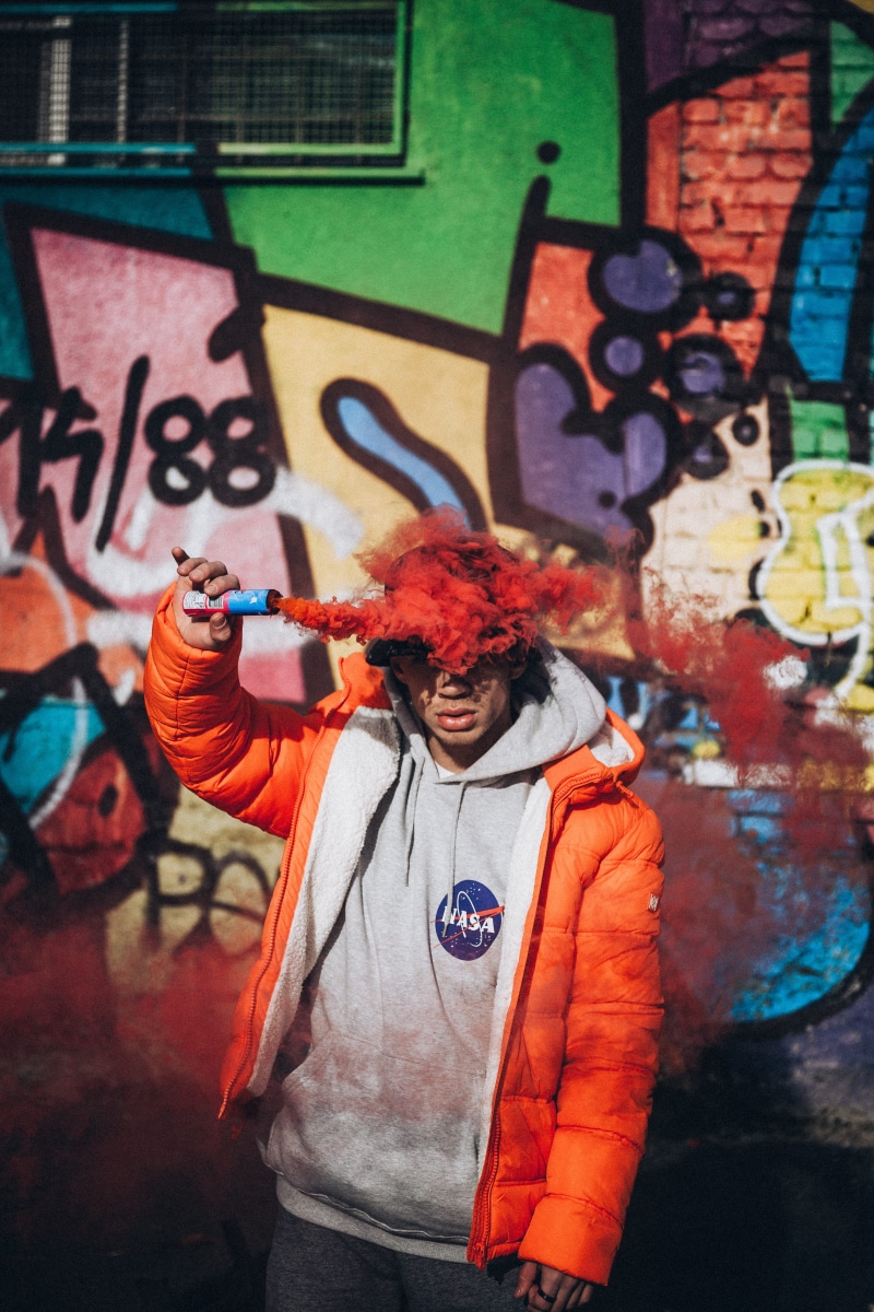 wall, clothing, architecture, jacket, abstract, casual clothing, graffiti, human, holding, hat, hair, smoke, urban, brick, red, multi colored, waist up