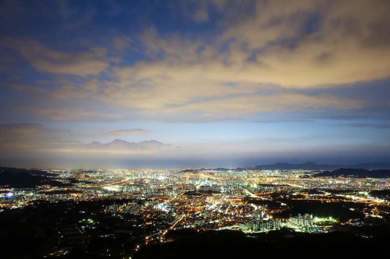 office building exterior, aerial view, cloud, mountain, outdoors, building exterior, high angle view, illuminated, sky, crowd, built structure, a night view of seoul