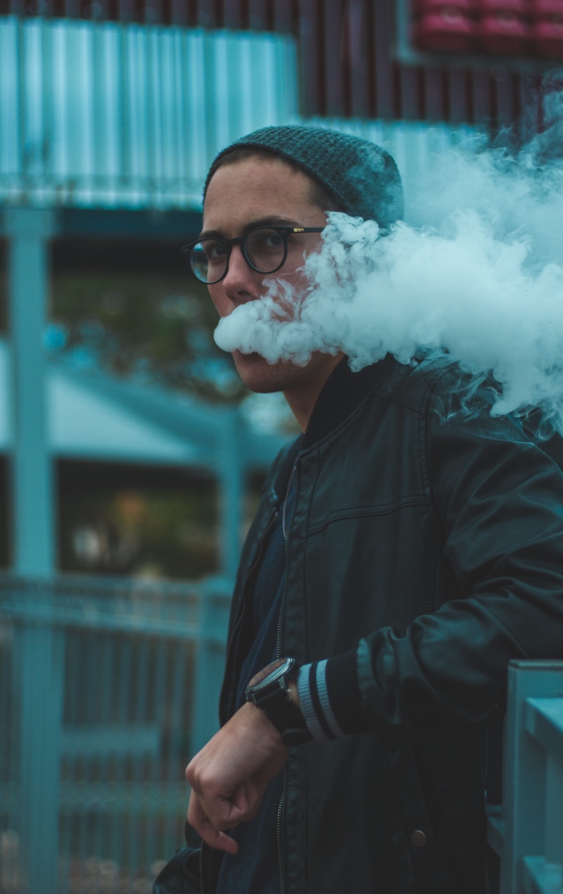leaning, standing, city, smoke - physical structure, one person, vaping, outdoors, young adult, eyeglasses, beard, waist up, smoke, coming, clothing, looking at camera